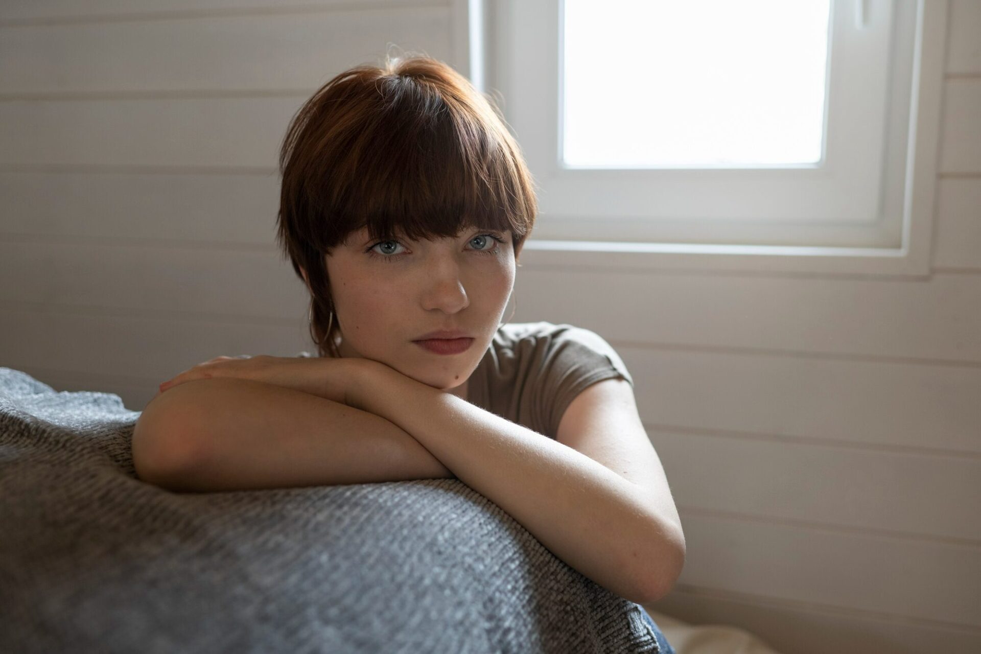 Young person resting their head on their arms, looking thoughtfully at the camera in a softly lit room