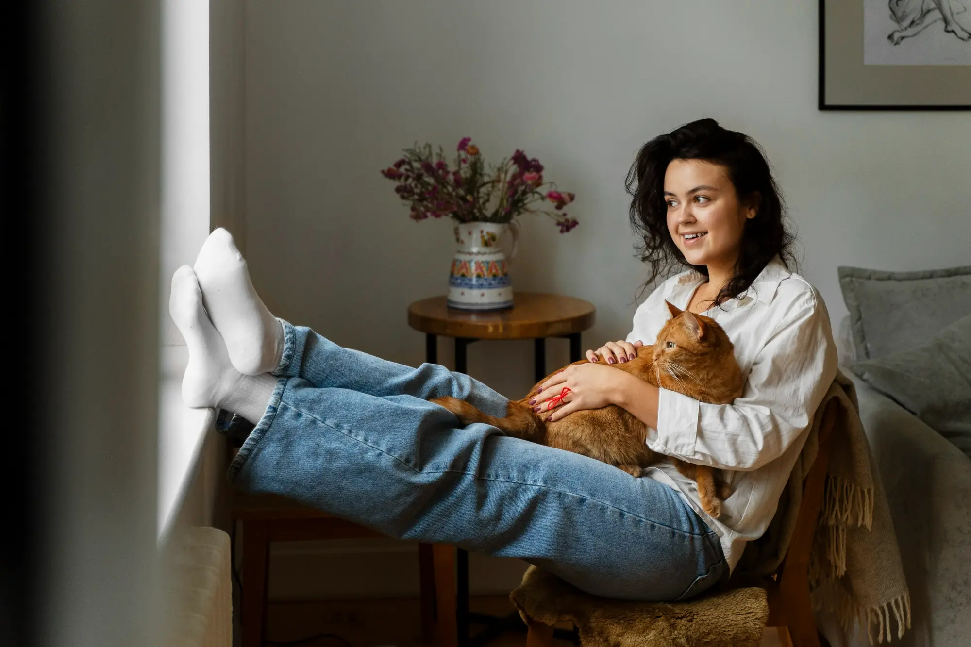 Woman relaxing at home with her feet up, holding an orange cat and looking out the window