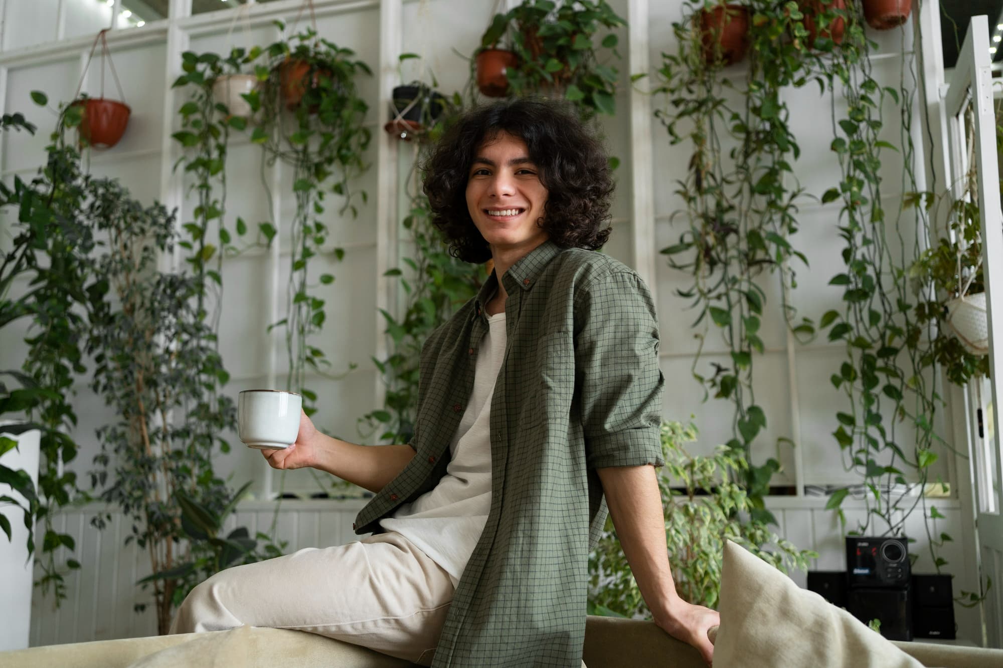 Young man smiling and holding a mug of coffee, surrounded by hanging plants at home
