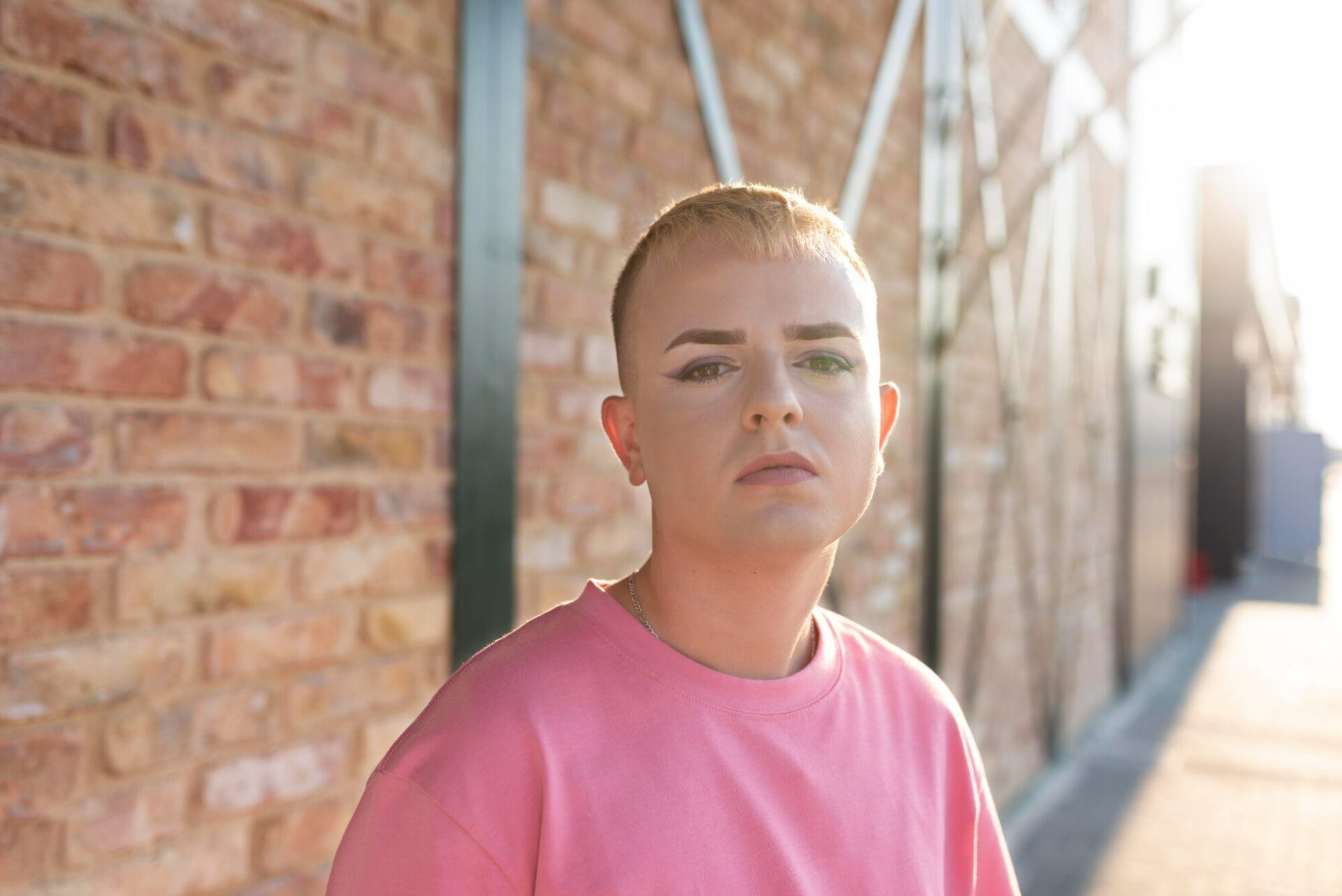 Young person with short bleached hair and makeup standing against a brick wall, looking directly at the camera